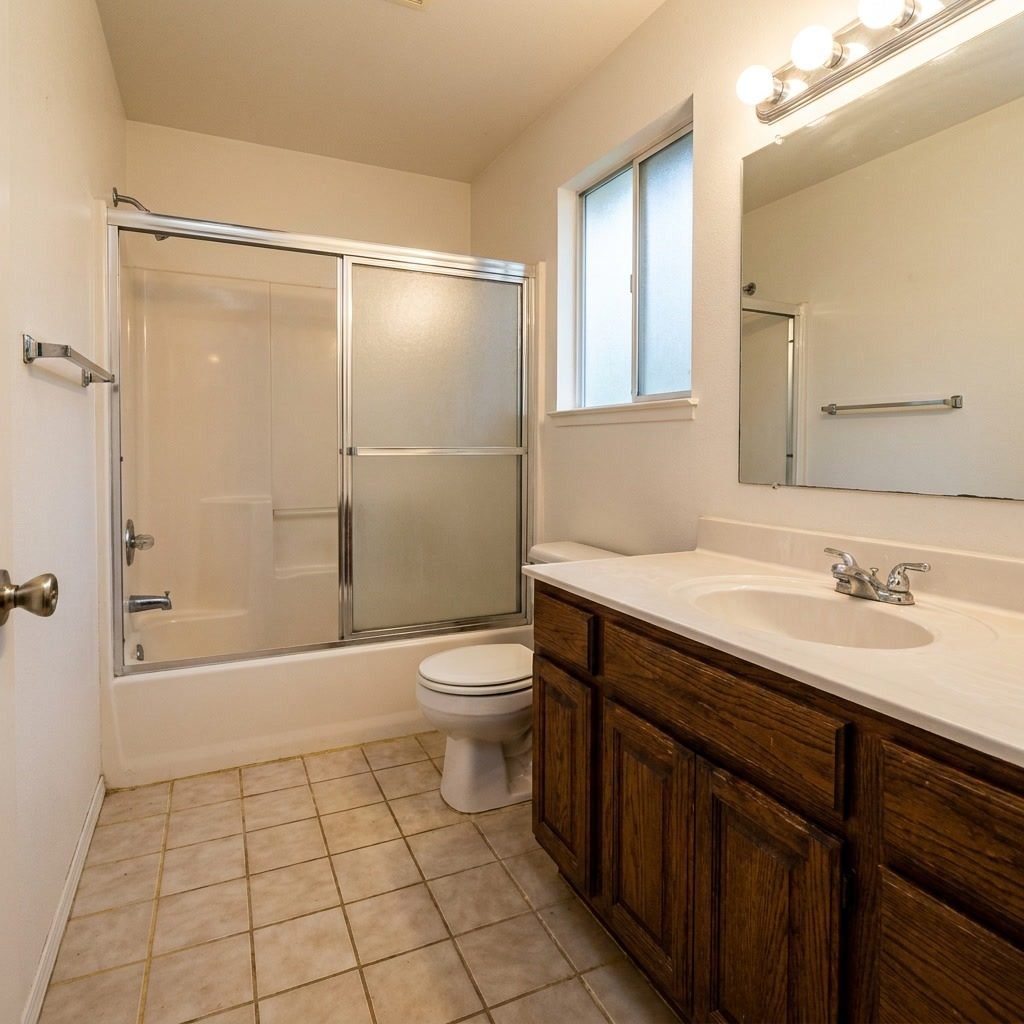 Before: Dated 1990s bathroom with chrome sliding glass tub-shower doors, beige ceramic tile floor, oak vanity, and old Hollywood light bar in Delray Beach, FL